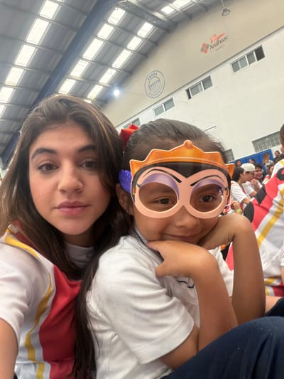 Two females are posing for a selfie. The younger female is wearing novelty eyewear, while the older female is wearing a red bow in her hair. Both appear to be wearing matching red and white sports jerseys. There are other individuals in the background, some of whom appear to be seated in bleachers. The location is likely an indoor gymnasium in Tlaxcalancingo, Mexico.