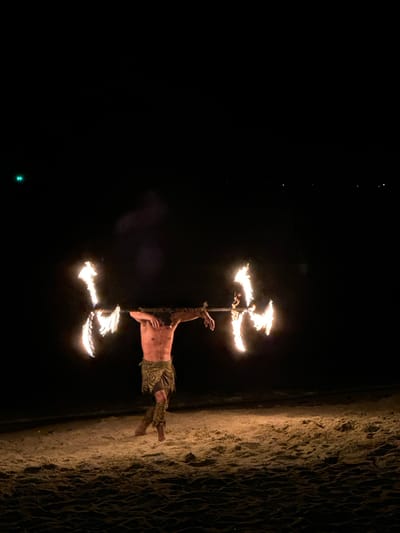 The image captures a nighttime scene on the beach of Hat Khai Muk, Thailand, where a man performs a fire dance. He is shirtless, wearing patterned pants, and holding a staff with lit flames at both ends. The ground is sandy, and the backdrop consists of a dark sky and the ocean.