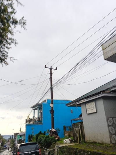 The image depicts an urban residential street scene in Sindangjaya, Indonesia, under a uniformly overcast sky. A prominent brown wooden utility pole stands in the center-midground, heavily burdened with a complex network of numerous black overhead electrical and communication cables that crisscross the frame. To the left of the utility pole, a two-story building painted bright blue features a second-floor balcony with a dark railing. To the right, a single-story building with light grey walls and a dark green corrugated metal roof is partially visible. A rectangular relief carving, displaying script consistent with Arabic calligraphy, is affixed to its exterior wall. In the lower left quadrant, the rear section of a dark-colored SUV is partially visible on what appears to be a wet asphalt road surface. Further down the street on the left, additional residential structures and indistinct smaller vehicles are present along a sloping incline. Green foliage from a tree is blurred in the upper left corner, and shrubs are visible in front of the blue building. A moss-covered concrete retaining wall occupies the immediate lower foreground, with minor debris scattered near the base of the utility pole. No clearly identifiable individuals are present.