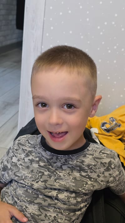 A young Caucasian boy with light brown hair and brown eyes is shown from the chest up, looking directly at the camera and smiling with his mouth open. He is wearing a camouflage t-shirt with a black collar. In the background, a yellow garment with a blue and white logo is visible, along with a wall with a subtle star pattern and part of a brick wall. The scene appears to be indoors, possibly in a home environment in Bulgakovo, Russia.