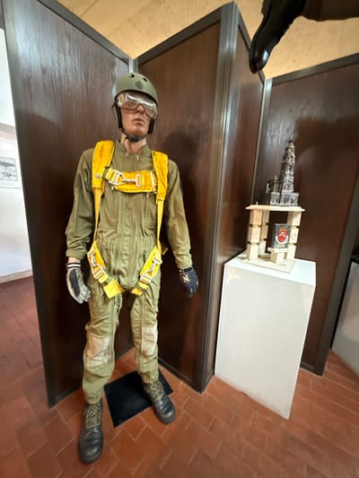 An indoor scene within a facility in Praia do Ribatejo, Portugal, depicts a full-sized mannequin dressed in military or paratrooper attire. The mannequin stands facing forward, wearing an olive green jumpsuit with a prominent yellow parachute harness featuring silver buckles and straps. A dark olive green helmet with clear goggles covers its head, and dark boots are on its feet. Its hands wear black and grey gloves, the right glove displaying a white "N" logo. The mannequin stands on a small black mat placed on a terracotta-tiled floor with a herringbone pattern.
To the right of the mannequin, a white rectangular pedestal supports a display. On top of this pedestal, a dark, intricate model of a tall, multi-tiered structure, possibly a church or tower, is presented. This model rests on a light-colored wooden base composed of four vertical posts and connecting elements, framing a small red and black crest or emblem.
The background consists of dark brown wooden paneling or doors. To the far left, a white wall with a framed picture is partially visible. In the upper right corner of the frame, a dark, upward-pointing object resembling the underside of a boot or shoe is partially visible, mounted to an unseen structure. The scene is evenly lit, suggesting an exhibition or museum environment.