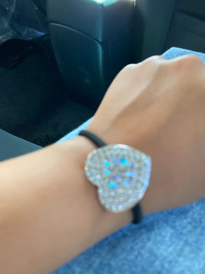 The image shows a close-up of a person's wrist wearing a black hair tie with a heart-shaped charm covered in rhinestones. The charm has iridescent blue and purple hues. The person is wearing a blue denim garment. The background is blurry but appears to be the interior of a vehicle, possibly a car, with dark seating and a mesh-like texture on what could be a floor mat. The specific location in Isyangulovo, Russia, cannot be determined from the visual information provided in the image.