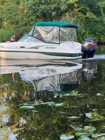 The image shows a white and green boat with a green canopy, labeled "Hurricane," with a "Suzuki Four Stroke" outboard motor, moving on a body of water, reflecting the surrounding trees. The boat has a partially transparent canopy covering the cabin area, and two individuals are partially visible inside. One person in the front appears to be a woman with her hair in a bun, wearing sunglasses. The boat is in motion, creating a wake. The water is calm and reflects the lush green foliage of trees that line the shore, as well as the boat itself. Lily pads are scattered on the surface of the water. The lighting suggests it is daytime, possibly late afternoon or early evening, with warm light illuminating the scene. The location is specified as Šyša, Lithuania.