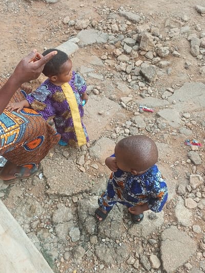 Two young children stand on a cracked concrete surface. One child wears a purple and gold patterned outfit with blue sandals. The other child wears a blue and white patterned outfit with black sandals. An adult wearing patterned clothing stands next to one child, with their hand near the child's head. Two small toy cars lie on the ground nearby.