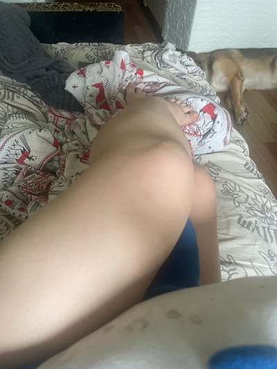 In the original image, the focus is on a person's legs, which are positioned on top of a patterned duvet. The duvet features a repetitive design of figures in red and black. The person's left foot is visible, and they appear to be lying down.
In the background, a dog is lying on the floor, facing away from the camera, its head and paws visible. The dog has brown and black fur. The floor appears to be made of wood. To the left of the dog, there's a wall with a textured surface.
The lighting suggests it could be daytime, but the exact time is not discernible. The scene appears to be indoors, likely a bedroom, given the presence of the duvet and the relaxed posture of both the person and the dog. There is no visible text in the image. The overall impression is one of a casual, domestic setting.