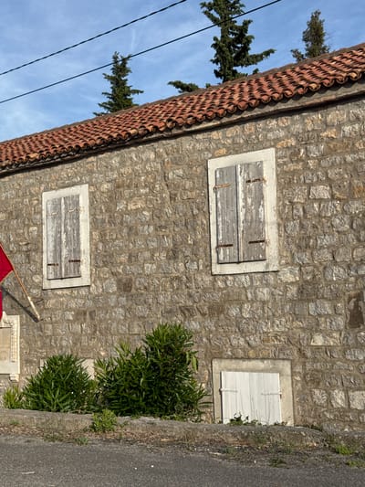 The image shows the facade of a stone building in Donji Katun, Montenegro, under a clear blue sky with thin clouds. The building features a terracotta tile roof and is constructed from irregular, light-colored stones. Two windows with weathered, grey wooden shutters are visible on the upper part of the facade. The left shutter is slightly ajar. Below the windows, a red flag on a white pole is partially visible on the left side of the image. To the right of the flag and below the left window, there is a light-colored stone architectural element. In the lower section of the image, a small, white-paneled door is set into the stone wall, and in front of the building, there are clusters of lush green bushes. A portion of an asphalt road and a concrete curb are visible in the foreground. The scene appears to be a static, outdoor view of residential architecture in a Mediterranean or Balkan setting.