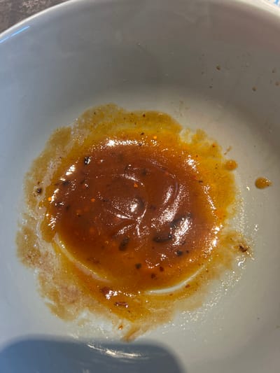 A close-up overhead shot shows the remnants of a reddish-orange sauce with visible chili flakes in a white ceramic bowl. The sauce is concentrated in the center of the bowl, with thinner streaks and droplets spreading outwards. The bowl itself appears to be part of a table setting. There are no people or recognizable landmarks of Leiden, Netherlands in the image.