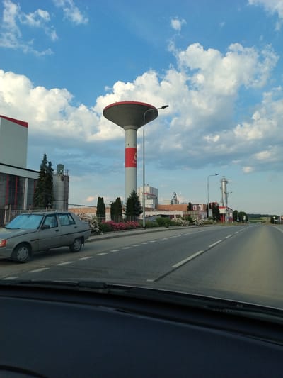 The image depicts a view from a vehicle on a road in Horažďovice, Czechia. On the left side of the road, a grey sedan is parked. Beyond the car, an industrial complex is visible, featuring various buildings, including a tall cylindrical water tower with a red top and a red circular logo. The complex also includes silos and smokestacks. The road is asphalt with white lane markings and is bordered by a sidewalk and a row of flowering bushes. In the background, trees and a green landscape with rolling hills are present under a blue sky with scattered white clouds. The scene appears to be captured during the daytime.
