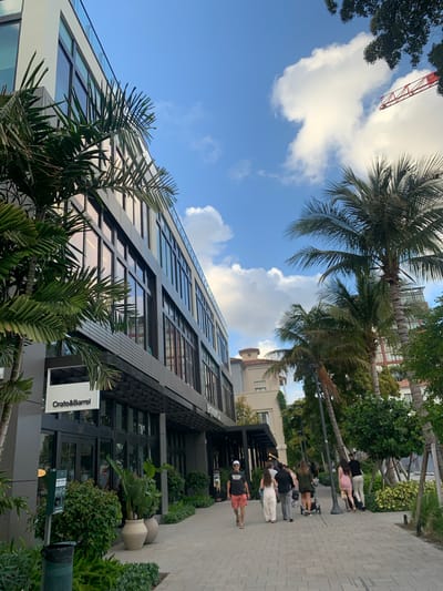 The image shows the Crate and Barrel store in West Palm Beach, Florida. The modern building is constructed with numerous glass windows. A sidewalk runs adjacent to the building, with several pedestrians walking along it. Palm trees and other greenery are present in the foreground and background. Above the building, the sky is blue with scattered clouds. A red construction crane is visible in the upper left of the frame.