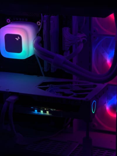 The image depicts the interior of a white computer tower case illuminated by colorful RGB lighting. A white CPU cooler with a glowing, multifaceted light effect is visible in the upper left corner, displaying shades of blue, green, purple, and pink. To its right are several white RAM modules, some labeled "FURY." A white GPU with the "GEFORCE RTX" logo visible is mounted horizontally in the lower half of the frame. This GPU also features RGB lighting, with a blue and green glowing outline visible on its side. Two large, white computer fans are situated on the right side of the case, emitting pink and blue light. The overall environment is dark, with the RGB lighting being the primary light source, creating a vibrant and high-tech aesthetic. There are no people or any indications of the city of Poritsy, Russia within the image.