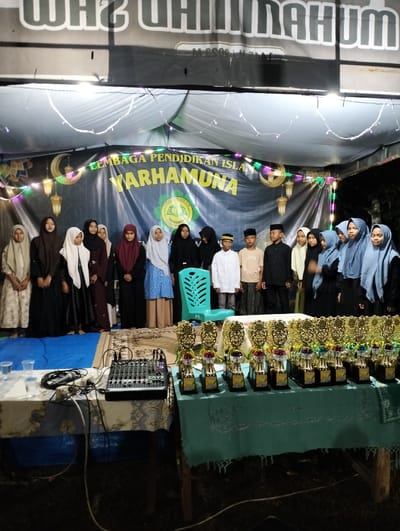 The image displays an outdoor or semi-outdoor nighttime event in Jambo Dalam, Indonesia. A group of approximately 17-18 children and adolescents are positioned in the mid-ground, standing in a line and facing the viewer. The group includes females wearing diverse colored hijabs and long garments, and several males wearing peci and long-sleeved shirts.
Behind the individuals, a dark blue banner serves as a backdrop, featuring the text "LEMBAGA PENDIDIKAN ISLAM YARHAMUNA" and a green circular emblem depicting an open book. The banner is adorned with decorative crescent moons and lantern cutouts along its top edge. Overhead, a white tarp canopy is illuminated by string lights. Above the canopy, a large black banner with partially visible reversed white text is present.
In the foreground, two wooden tables are positioned. The left table, draped with a white and blue patterned cloth, holds a black audio mixer, a wired microphone, and several clear plastic cups, some containing liquid. The right table, covered with a light green tablecloth, displays at least 30 gold-colored trophies with multi-colored bases, arranged in multiple rows. A single light-blue plastic chair is visible behind the left table, positioned to the right of the standing group. The ground area in front of the group is covered by a blue tarp and a patterned rug.