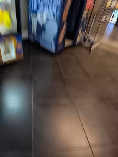 The image depicts an indoor commercial or public space, possibly a corridor or a section of a store, in Agios Dimitrios, Greece. The perspective is angled downwards, focusing on a dark brown or grey tiled floor with prominent grout lines.

On the far left, a bright yellow item is partially visible within a transparent display unit, likely a vending machine. Below it, a smaller object encased in yellow and blue packaging rests on the floor.

A large, blue display stand or vending machine stands prominently in the center-left. It features a graphic of a light blue human silhouette, depicting a head and shoulders with a speech bubble nearby. Above this graphic, white text is visible against the blue background. Although blurred, phrases like "STOP..." and "TO THE CALENDAR" are partially discernible, suggesting a promotional or informational message. To the right of this blue unit, vertical strips of reddish-brown elements, possibly snack packaging, are visible.

Further to the right, a metallic structure, resembling a railing or part of another display, extends into the background. The background beyond these structures appears to be a brightly lit interior space.

The image is characterized by significant motion blur, especially in the foreground and towards the right, contributing to a sense of movement or low-light conditions during capture. No individuals are present, and no specific activities are occurring within the frame. The lighting suggests artificial indoor illumination.
