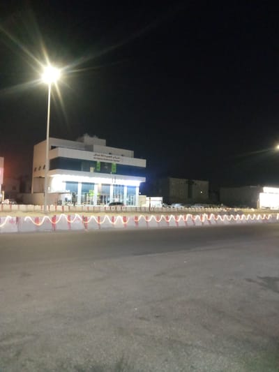The image depicts a street scene at night in Al Khobar, Saudi Arabia. A three-story white building, identified by signage in Arabic and English as "Al-Habra Business Center," is brightly lit. A tall street lamp stands adjacent to the building, casting a glare. In the foreground, a concrete barrier separates the building from the road. The barrier is decorated with red and white reflective tape. The road surface is dark gray. A row of buildings can be seen in the background. A few cars are parked near the business center.