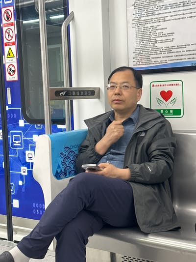 A middle-aged Asian man wearing glasses, a blue patterned shirt, and a dark gray jacket is sitting on a blue and white seat inside a train. The train car is decorated with a blue graphic pattern. A silver handrail is visible next to the window. A sign indicating courtesy seating and safety rules are mounted on the wall. Additional informational signs are visible above the man's head in Chinese. He appears to be holding a mobile phone in his hand. The scene suggests he's riding the Jiangtiao line, as indicated by signage.