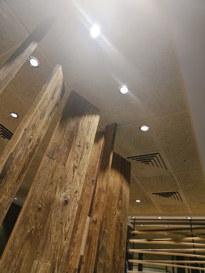 An interior shot looking up at a ceiling. The ceiling is tiled with a textured material and has several recessed lights. A large wooden beam structure is prominent in the foreground, angled upwards and to the right. A ventilation grate is visible on the ceiling. The lighting is bright, with some glare from the fixtures. The overall aesthetic is rustic and modern.