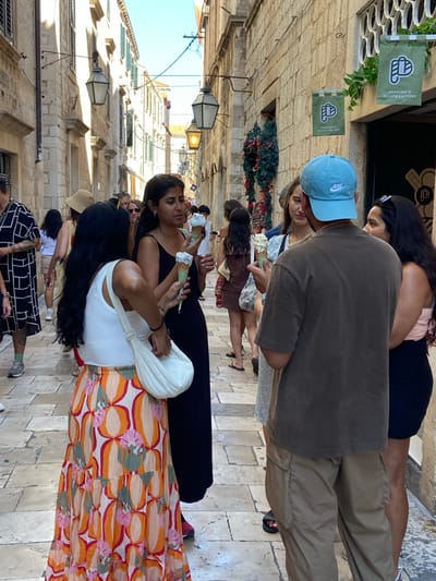 A group of people are gathered on a cobblestone street in Dubrovnik, Croatia, with the iconic stone buildings and lamps lining the narrow passage. In the foreground, a woman with long black hair, wearing a white tank top and a vibrant, floral-patterned skirt, holds an ice cream cone. She is interacting with a man wearing a blue Nike baseball cap and a brown t-shirt, who is also holding an ice cream. To their right, another woman in a black dress holds two ice cream cones. Behind them, several other individuals are visible, some holding ice cream as well, suggesting a shared experience of enjoying a sweet treat in the historic city. Banners for "Peppino's Gelato Factory" hang on the wall of a building, identifying the establishment. The scene captures a candid moment of leisure and enjoyment amidst the charming ambiance of Dubrovnik's old town.