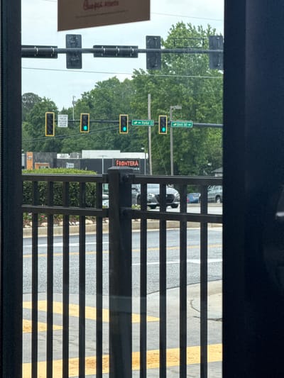 The image shows an exterior view through a window in Stone Mountain, Georgia, USA. A pole supports street signs that read "Parker Ct" and "Davis Rd". Traffic lights are on the pole with a green light illuminated. A sign reads "LEFT TURN, YIELD ON FLASHING YELLOW ARROW" and a business sign reads "FRONTERA KITCHEN". A road and yellow median can be seen, and the bars of a railing are visible. Cars are parked along the street.