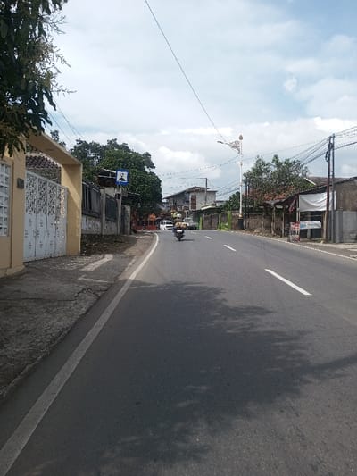 The image depicts a daytime scene on an asphalt road in the city of Chhenangri_Rishor, Bhutan, under a partially cloudy sky. The road, marked with a solid white line on the left shoulder and a dashed white line indicating a lane, extends into the distance. Tree shadows are cast upon the road surface.
On the left, a cream-colored building with a white ornamental metal gate stands beside a cracked concrete sidewalk. Further down on the left, a dark fence line runs parallel to the road, with a blue rectangular warning sign featuring a white pictogram visible on a pole. Beyond these, green trees and additional buildings line the street.
On the right side of the road, a utilitarian brown-toned building with a white tarp canopy and a sign displaying "BUHA" and other text is visible. Utility poles with a dense network of overhead wires are present along this side. A decorative street lamp post with a gold-colored finial stands prominently in the mid-distance on the right.
One individual is operating a dark-colored motorcycle, equipped with a helmet and carrying a large bundled load on its rear, traveling away from the viewer in the road's center-right lane. A white van or small bus is also visible in the far distance, moving in the same direction.