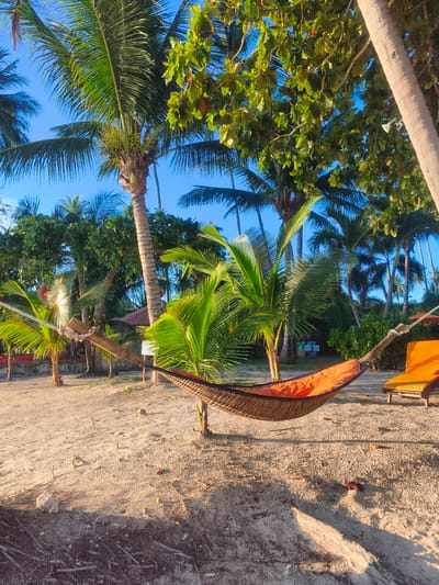 The image depicts a serene beach scene in Baan Lipa Noi, Thailand. In the foreground, a woven hammock with an orange cushion is suspended between two palm trees, inviting relaxation. The sandy ground is littered with fallen leaves and small branches, hinting at a natural beach environment. In the background, lush green palm trees and other tropical foliage dominate the landscape, with a clear blue sky overhead. To the right of the hammock, a bright orange lounge chair is visible, suggesting a resort or private beach setting. The overall impression is one of tranquility and tropical paradise.