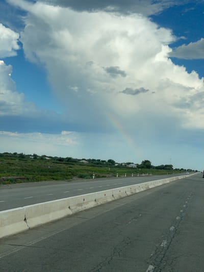 The image depicts a roadside scene in Qtay, Uzbekistan, under a cloudy sky with a hint of a rainbow. The perspective is from ground level, looking down a paved road that stretches into the distance. The road is cracked and weathered, with dashed white lines marking lanes. A concrete barrier with red markers separates the road from a grassy verge.
The verge is covered in green vegetation, with a line of trees further in the background. A few small structures are visible among the trees, possibly houses. The sky is filled with large, white cumulus clouds, with patches of blue sky peeking through. A faint rainbow arc is visible in the hazy sky. The overall impression is of a rural or semi-rural setting on an overcast day, perhaps after a rain shower. There is no visible text in the image. The time of day appears to be daytime, given the lighting.