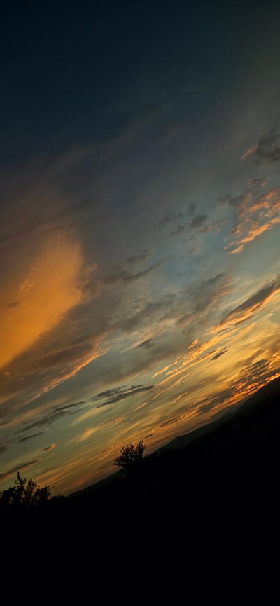 A photograph captures a dramatic sunset over a silhouetted landscape. The sky is a gradient of deep blue at the top, transitioning through shades of gray, teal, and finally a vibrant orange and yellow near the horizon. Wispy clouds are scattered across the sky, illuminated by the setting sun, creating streaks of light and shadow. In the foreground, the land is entirely black, obscuring any details of terrain or vegetation, except for a few dark shapes that might be trees. The overall impression is one of a vast and awe-inspiring natural spectacle, with the earth appearing as a dark, indistinct mass against the vibrant sky. The location is Momin Prohod, Bulgaria.