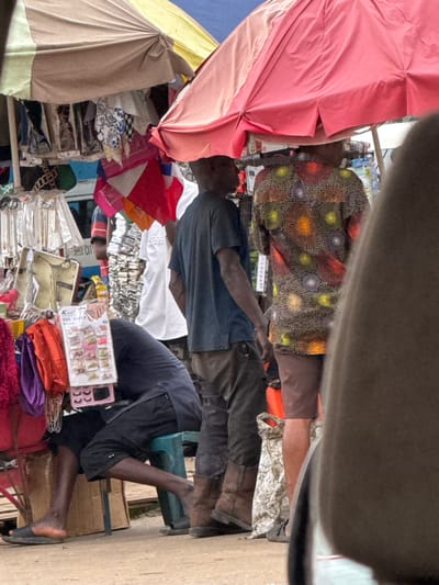 This is a medium-shot, eye-level photograph taken outdoors, depicting a bustling street market scene in Orji, Nigeria. The scene is dominated by colorful market stalls covered by large umbrellas. In the foreground, a person is seated on a green plastic stool, wearing a dark t-shirt and black shorts, with their head bent down as if attending to something on a display stand in front of them. The display stand holds various items, including a packet labeled "Toe Nails". Next to the seated person, a man in a dark t-shirt and brown pants stands, his arm partially visible. Further back, another person is visible wearing a vibrant, patterned shirt with circular designs in various colors. In the background, more stalls and people are present, indicating a lively market atmosphere. The ground appears to be a dirt or paved surface, typical of a market area. The overall impression is one of commerce and daily life unfolding in a Nigerian street market.