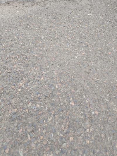 A close-up, eye-level shot depicts a rough asphalt surface, likely a road or path, in Stepnoe, Russia. The texture is uneven, composed of small grey and brown stones embedded in a dark grey binder. A few dry grass stalks are visible, scattered across the surface, suggesting an outdoor, possibly unkempt area. The lighting is diffused, indicating an overcast day or shade. There are no people or animals present, and no discernable objects beyond the natural composition of the asphalt and vegetation.