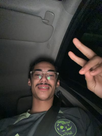 A young man in glasses smiles, showing braces. He makes a peace sign above his head. He is inside a car, facing the camera at an angle. The interior is visible, including the sun visor and roof. He wears a grey jersey with a patch that looks like a soccer ball surrounded by script. A seatbelt crosses his shoulder. The photo appears to be taken at night, indicated by the darkness in the background.