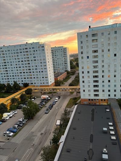 The image depicts a residential area in Solna, Sweden, with several tall, light-colored apartment buildings. The sky is filled with dramatic clouds, painted with hues of orange, pink, and yellow, indicative of a sunset or sunrise. In the foreground, the dark, flat roof of a lower building is visible, featuring various vents and skylights. Below, a street lined with parked cars is seen, with a white van bearing the "Best" logo parked among them. Trees and greenery are interspersed between the buildings and along the street. A pedestrian walkway or bridge connects two of the apartment buildings. The apartment windows show varying degrees of illumination, suggesting occupancy. The overall scene is a view from an elevated perspective, looking down on a functional urban environment during twilight.