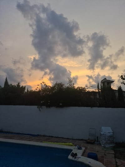 The scene depicts a residential outdoor area in El Campello, Spain, at dusk or dawn, characterized by a dramatic sky. The upper half of the image features an overcast sky with irregularly shaped dark grey clouds, particularly prominent in the upper left quadrant. Warm orange and yellow hues are visible along the horizon line, indicative of low sun illumination.

The lower half of the image presents a swimming pool in the foreground, with blue water filling the basin. The pool perimeter features light-colored coping tiles. On the coping to the right, a pair of dark sandals and dark swimming goggles are placed. Adjacent to the pool on a tiled patio, a yellow inflatable pool accessory is visible, along with a blue plastic bucket and a green garden hose.

A solid, light-colored concrete or stucco wall horizontally bisects the mid-ground, acting as a boundary. To the right of the pool and against this wall, a metal utility cart or hand truck is present, and next to it, a large object covered in white sheeting.

Above the wall, the background consists of dense, dark green foliage, primarily trees and bushes, silhouetted against the sky. Clusters of small white flowers are visible among the foliage directly above the wall. To the far right, a partial view of a building structure with a prominent, square-shaped chimney or vent stack is discernible, partially obscured by vegetation. Tall, slender evergreen trees, possibly cypresses, pierce the skyline on both sides of the background. No individuals or active interactions are present in the image.