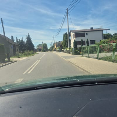 This image depicts a street scene in Mnichowice, Poland. The perspective is from inside a vehicle, looking out through the side window. The scene is oriented sideways, with the sky appearing on the left and the road on the right.

The sky is a clear blue with wispy white clouds, suggesting a pleasant day, likely late morning or early afternoon due to the bright sunlight. A road with double white lines runs through the scene, bordered by a paved sidewalk on the left.

Along the sidewalk, there are houses and vegetation. On the left side of the road, there are several houses, some appearing modern with white facades and dark roofs, others with more traditional architecture. Trees, including tall evergreen trees and deciduous trees with green foliage, are interspersed with the houses. There are also fences, one of which is a green metal mesh fence with flowering plants behind it. Power lines are visible strung between utility poles, indicating the presence of infrastructure.

In the distance, the spire of a church or similar tall building can be seen peeking above the trees. Some street signs are also visible near the road, though the text is not legible at this resolution. The overall impression is a quiet, residential street in a Polish village or town on a sunny day. There are no people visible in the image, and the activity appears to be simply the passage through the area. The image is captured in a sideways orientation, which needs to be corrected for proper viewing.
