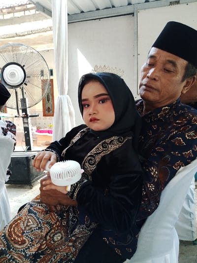 A man and a young girl are seated together in an open-air or semi-enclosed event space in Juwana, Indonesia. The man, a middle-aged or elderly male with a dark complexion, wears a dark batik shirt and a black peci. He is seated on a white plastic chair, leaning back with his eyes partially closed, looking towards his right. The girl, appearing to be a child, sits on the man's lap, wearing a black long-sleeved dress with gold embroidery, a black hijab, and a silver tiara. Her face is heavily made up with pink eyeshadow, dark eyeliner, blush, and red lipstick. She holds a white portable handheld fan with her right hand and looks directly at the camera with a neutral expression. In the background, a large industrial standing fan is visible on the left, and white decorative fabric drapes are tied around a pole. A corrugated metal roof structure is visible above, and an open doorway or window with a decorative frame is partially visible in the distance. A black equipment case on wheels is also present in the left background.