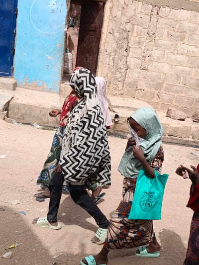 An outdoor scene in Zango, Nigeria, depicts four individuals walking on a dirt path alongside weathered buildings. In the foreground right, a young person, likely a child, wearing a light green hijab and a patterned dress, holds a clear plastic bag to their mouth. A turquoise cloth bag with "AISHA" printed on it hangs from their left shoulder. Directly behind them, an individual wearing a black and white zigzag patterned outer garment over a colorful dress and black trousers walks left-to-right. To the left of this individual, another person, partially obscured, wears a red and white patterned garment over a blue and green patterned dress. A fourth person, mostly hidden behind the zigzag-patterned individual, wears a light pink head covering. All visible individuals wear slide sandals. The ground is uneven, dusty, and littered with small debris. The background features a bright blue wall or gate on the far left, followed by a textured light brown mud or concrete block wall with a dark brown wooden door centrally located. Another section of the textured block wall with faint markings is visible on the right.