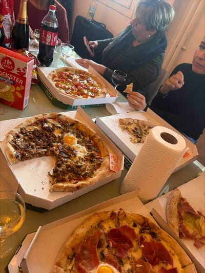 An indoor scene in Cagnes-sur-Mer, France, depicts a casual meal gathering around a light-colored table. Four individuals are partially visible. In the left background, a person in a red top is near two uncorked champagne bottles. In the center-right background, a woman with short, light-brown hair and glasses, wearing a dark top, holds a white smartphone with her left hand. To her right, a woman with dark hair and a black top holds a piece of pizza crust in her left hand, positioned near the right edge of the frame.
The table is laden with multiple open pizza boxes. One pizza features ground meat and two sunny-side-up eggs, with one slice removed. Another pizza displays red sauce, yellow vegetables, and red toppings. A third pizza contains prosciutto and what appears to be a sunny-side-up egg. A partial pizza with mushrooms is visible in another box, along with a solitary slice in an adjacent box. A full roll of white paper towels stands upright on the table. Beverages include a Coca-Cola Zero Sugar bottle, a box of "Carrefour Classic Snackster Original" crisps, and several glasses, including two with red wine and one with a clear, sparkling liquid. Small condiment packets are present in one pizza box. The background shows light-colored walls and a dark bag.