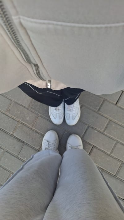 This image shows a ground-level perspective of two pairs of feet wearing white sneakers, positioned on a paved walkway. The feet in the foreground are clad in light gray sweatpants, while the feet further back are wearing dark pants. A portion of a light-colored jacket with a visible zipper is in the upper part of the frame. The walkway appears to be constructed of rectangular gray paving stones. The scene is outdoors, as indicated by the natural lighting and the presence of pavement. The location is identified as Stowbtsy, Belarus, though no specific landmarks are visible to corroborate this.