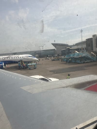 The image depicts an airport tarmac scene, likely in Gwaska, Nigeria, given the context. In the foreground, a portion of an airplane wing is visible, extending from the bottom left to the bottom right of the frame. The wing is a light gray color with some metallic paneling and rivets. Beyond the wing, an airplane is parked. It's a narrow-body jet with a white fuselage and blue and red accents. The plane appears to be undergoing some form of maintenance or servicing, as evidenced by a vehicle with a crane-like apparatus positioned near its side, and several people in yellow vests and hard hats are gathered around it.
Further in the background, there are airport terminal buildings with a distinctive architectural style. A set of mobile passenger stairs, colored light blue, is parked near another aircraft, also with people in high-visibility vests visible on and around it. The tarmac is paved and appears to be a busy operational area. The sky above is overcast with some clouds, suggesting a daytime scene. The overall impression is one of an active airport environment, possibly during boarding or deplaning procedures, or ongoing aircraft service.