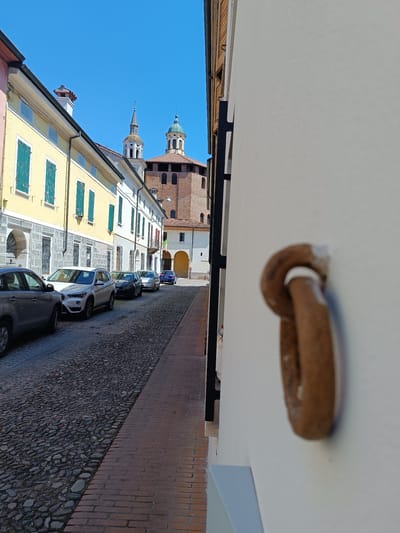 A cobblestone street in Mezzana Sant'Antonio, Italy, lined with parked cars and buildings with colorful facades and green shutters. In the distance, the bell tower and dome of the Sanctuary of the Madonna della Fontana are visible. An old, rusty metal ring is affixed to a white wall in the foreground.