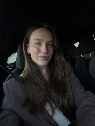 A woman is seated in the driver's seat of a car, wearing a brown sweatshirt and a seatbelt with a white strap. She is looking at the camera with a slight smile. Her hair is long and brown. The background is blurred, but shows a glimpse of greenery through the car window, suggesting a location in a natural environment. This is Grabovica, Montenegro.
