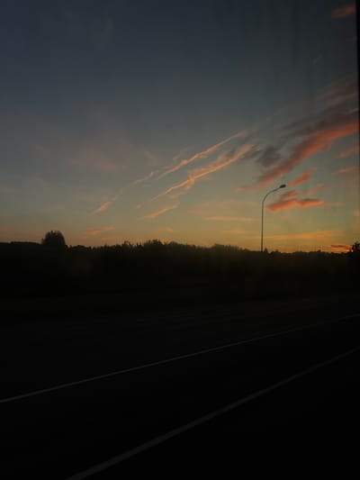 This image depicts a twilight scene, likely in Tri-Izby, Russia, with a dark silhouette of trees and a road in the foreground. The sky is a gradient of deep blue at the top, transitioning to orange and yellow hues near the horizon, with wispy, pink-tinged clouds streaking across. A solitary street lamp stands tall against the darkening sky, its light extinguished. The road, which appears to be a highway, is dark and marked with white lines, suggesting it is either a highway or a significant road. The overall atmosphere is serene and slightly melancholic, characteristic of a rural or suburban landscape at dusk.