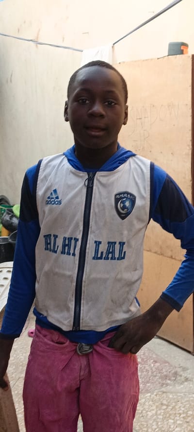 A young boy stands indoors wearing a white and blue Adidas jacket with "Al Hilal" printed on it and the Al Hilal FC logo. He also wears pink pants with a knotted rope or belt. The background appears to be a simple courtyard with light-colored walls. Graffiti is visible on the wall behind him.