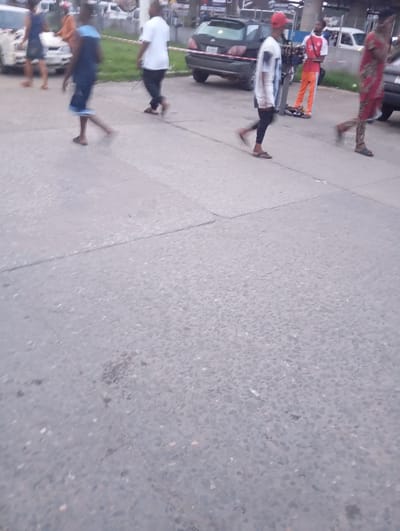 A group of people are walking on a paved street in Port Harcourt, Nigeria. In the foreground, the grey asphalt is cracked in several places. In the background, several cars are parked or driving, including a white van, a black SUV, and a lighter colored sedan. There are also some people in the background, including a woman in a blue dress and a man in a white shirt. The overall impression is of a busy urban environment.