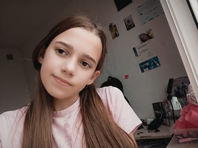 The image shows a medium shot of a young Caucasian female with long brown hair and fair skin, wearing a light pink t-shirt. She is looking towards the camera with a neutral expression. The background is a cluttered desk in what appears to be a bedroom. On the desk, there are various items including a white lamp, a bottle of micellar water, a phone charger, a black electronic device, and a clear plastic container filled with pink hair curlers. The wall behind the desk is adorned with several pictures and certificates. The overall lighting suggests an indoor setting, possibly during the daytime given the natural light coming from the right side of the frame. The location is identified as Radzymin, Poland, implying the scene is within a residential property in that city.