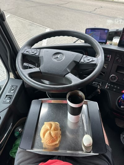 In the cab of a Mercedes-Benz truck in Schipkau, Germany, a stainless steel tray rests on the driver's lap. On the tray is a sandwich on a kaiser roll, a cup of coffee in a black and white thermal mug, and a hard-boiled egg on a spoon. To the left of the tray, a plastic water bottle and a green plastic bag are visible in a storage compartment. The steering wheel of the truck, with the Mercedes-Benz logo, is in the foreground, and a GPS device is mounted on the dashboard. The driver's legs are visible, wearing grey sweatpants and a red t-shirt. The exterior of the truck is visible through the windshield, showing a grey paved area with white markings.