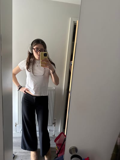 A young woman is captured in a mirror selfie, standing upright in what appears to be an indoor room in Hull, United Kingdom. She has long, dark brown hair, wears reddish-brown framed glasses, and white over-ear headphones. Her expression is neutral, with a slight pout as she looks towards her phone. She is dressed in a plain white short-sleeved t-shirt, black wide-leg trousers, and light brown slip-on shoes. On her wrists, she wears a beaded bracelet on her left and a multi-strand fabric bracelet on her right. A thin necklace is also visible around her neck.

She holds a light yellow smartphone, featuring a black camera lens and a textured case, in her right hand to take the photo. Her left hand is placed on her hip.

The background shows a minimalist room with light-colored, possibly white or off-white, walls and a light-toned floor, which could be laminate or tiled. Directly behind her is a white horizontal radiator, indicating a heating system, with visible pipes underneath. A subtle light fixture, possibly an LED strip light, runs along the top edge of the wall above the mirror, providing illumination. To the right, on the floor, parts of a red bag and possibly another blue item are visible. No explicit text is discernible within the image. The setting suggests a casual, everyday moment indoors.