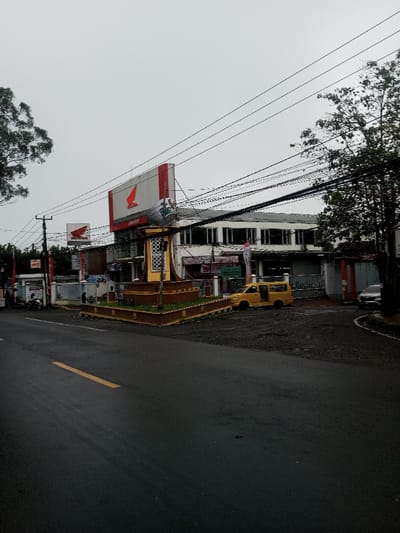 This image depicts a commercial street scene in Chagzam_Pam, Bhutan, under an overcast sky. In the foreground, an asphalt road features a single yellow dashed center line. Overlapping the road and the mid-ground, numerous dark utility lines and cables crisscross the upper half of the frame.
In the mid-ground, a central, square-shaped, multi-tiered monument or planter box with yellow and brown coloration is situated on a paved area, containing greenery and featuring decorative white patterns on its central pillar. Directly behind this monument, a large red and white billboard prominently displays the "Honda" logo, with partially visible text below that reads "CONCES...". To the left of this main billboard, a smaller "Honda" sign is also visible.
Behind the signage and monument stands a multi-story commercial building with a light-colored facade and multiple dark-framed windows across its upper levels. The ground level of this building includes an entrance with a fence or gate. To the left of the monument, a white scooter or motorcycle is parked near a utility pole. To the right of the monument, a yellow van, possibly a Suzuki Carry, with its side door open or missing, is parked on an unpaved section of ground. A dark-colored SUV is partially visible, parked further to the right, near the edge of the frame. Deciduous trees are present on both the far left and right sides of the image. Several red and white banners or flags are attached to poles and fences in front of the buildings. No clearly identifiable individuals are engaged in distinct actions.