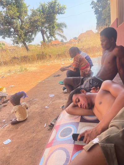 An outdoor scene in Oturkpo, Nigeria, depicts four individuals in various states of repose under bright daylight. In the foreground, a shirtless male with facial hair lies on his left side on a multi-colored patterned mat, head resting on his left arm, with eyes closed. His right hand holds a black smartphone. He wears khaki shorts. Adjacent to him, a shirtless male sits against a reddish-brown wall, wearing dark shorts with a white drawstring, facing left. Behind him, another male is seated, wearing a striped orange, yellow, and blue shirt and dark pants, also facing left. Further to the left on the reddish-brown dirt ground, a small child is seated, facing away, wearing a light-colored, possibly checkered, shirt and a blue hair accessory. The ground transitions from a paved or tiled surface where the adults are seated to a dirt area littered with plastic bags and wrappers. Beyond the immediate foreground, dry, tall grass and shrubs line the background, punctuated by a large leafy green tree and a palm tree under a clear sky. Distant structures with distinct roofing are visible through the sparse vegetation. A sharp shadow line diagonally crosses the dirt ground, separating a shaded area from sunlit terrain.