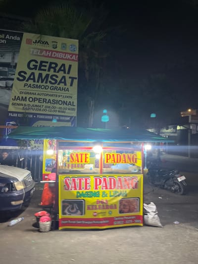 The image depicts a night scene on a street in Tangerang, Indonesia, near the Graha Autoparts Station. A prominent yellow banner advertises "GERAI SAMSA" which translates to "SAMSAT OUTLET". This outlet appears to be a government service center for vehicle registration and tax payments. The banner indicates the outlet is open, with operating hours listed for weekdays and Saturdays.
In the foreground, a food stall is set up under a green awning, selling "SATE PADANG" (Padang satay). The stall displays images of the food and contact information for orders. A blue car is parked to the left of the food stall, and a black motorcycle is parked to the right. The street appears to be asphalt. The overall atmosphere is that of a bustling urban environment at night, with street vendors and public service access.