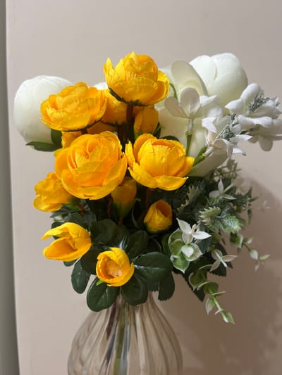 A bouquet of artificial flowers is displayed in a ribbed, translucent glass vase. The bouquet is comprised of several bright yellow rose-like flowers, two larger white flowers that appear to be peonies, and smaller white flowers with delicate petals, interspersed with green foliage and eucalyptus-like greenery. The background is a plain, light-colored wall. The image is a close-up shot of the floral arrangement, with no people or specific city locations depicted.