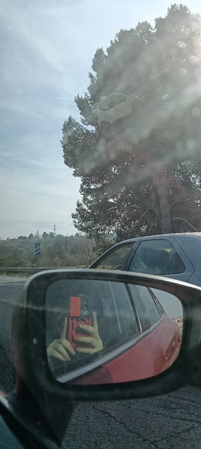 The image depicts a roadside scene observed from the interior of a red vehicle, likely in Sant Quirze del Vallès, Spain. In the foreground, the vehicle's driver's side mirror reflects the hands of a person holding a red smartphone, encased in a matching red protective cover, positioned for photography. The reflection also shows part of the red vehicle's door and asphalt below.
Outside the vehicle, to the immediate right, the upper section of a dark blue or gray hatchback is visible. A large, dense evergreen tree with dark foliage dominates the upper right and central parts of the frame. The sky, visible above and through the tree, is light blue with sparse cloud cover, featuring a thin white aircraft contrail in the upper left quadrant. A reflection of a light-colored SUV is superimposed over the tree and sky in the upper windshield area.
To the left, a metal guardrail separates the roadway from an elevated hillside characterized by sparse, dry vegetation. A blue rectangular road sign displaying white text, indicating a distance possibly as "700 m," is visible on this hillside. A tall electricity pylon stands on the distant horizon to the left, above the hillside. The ground appears to be paved with asphalt. The scene is illuminated by natural daylight, with a lens flare indicating the sun's position off-frame to the upper right.