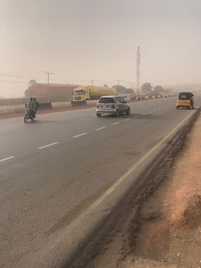 An asphalt road curves slightly with a white dividing line. A motorcycle with two people is in the foreground, followed by a silver car and a yellow truck with “Green Ville Lux” on it. Further ahead is a yellow tricycle. A cell phone tower rises beside the truck and car. Hazy or foggy conditions obscure the distant background. The shoulder of the road is dirt and gravel.